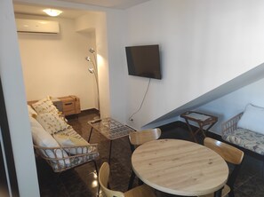 A living room with a sofa, chairs, a table, a TV mounted on the wall, and an air conditioning unit.