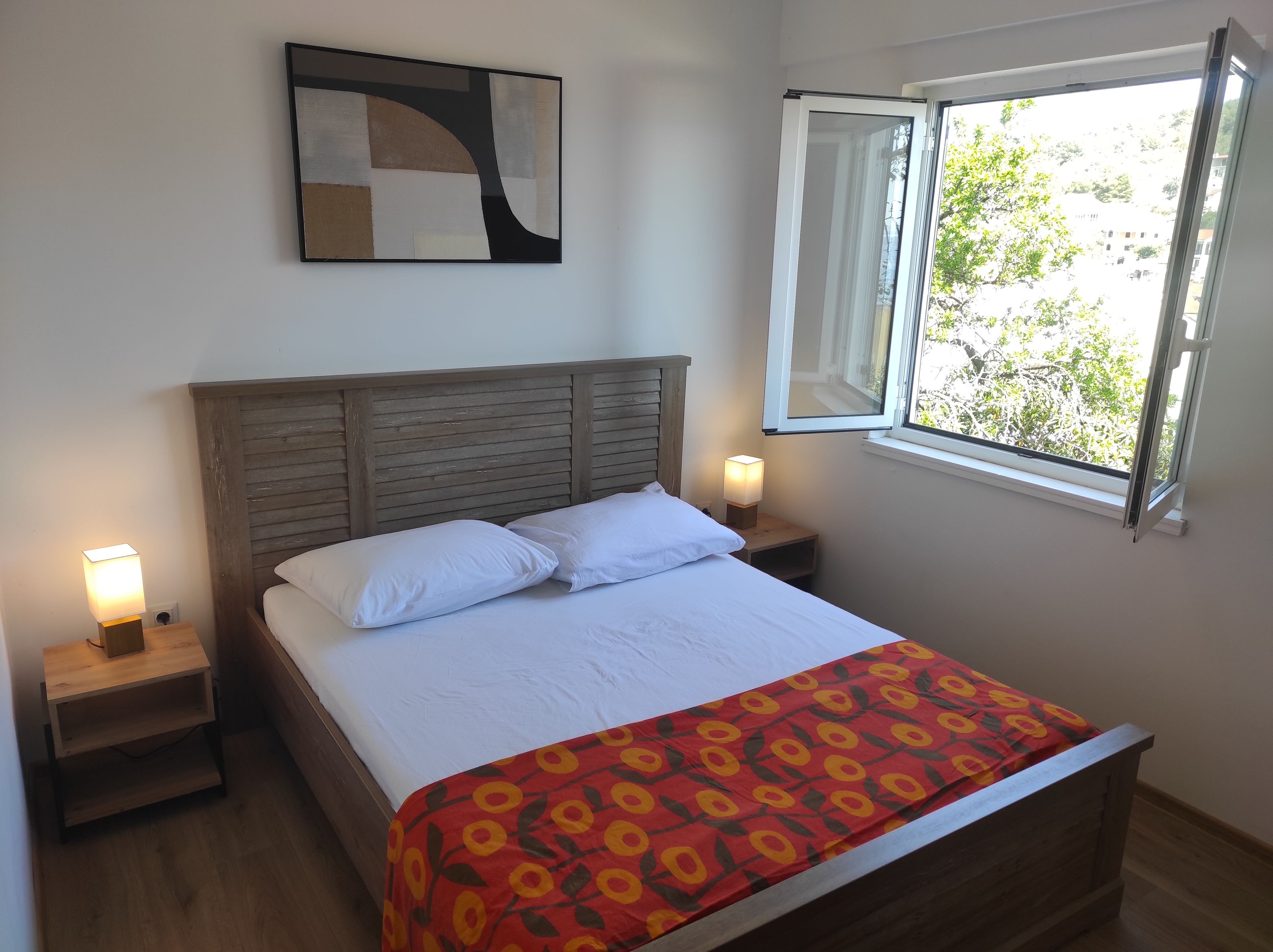 A bed with a patterned bedspread, a wooden headboard, two bedside lamps, and a window with an open view.