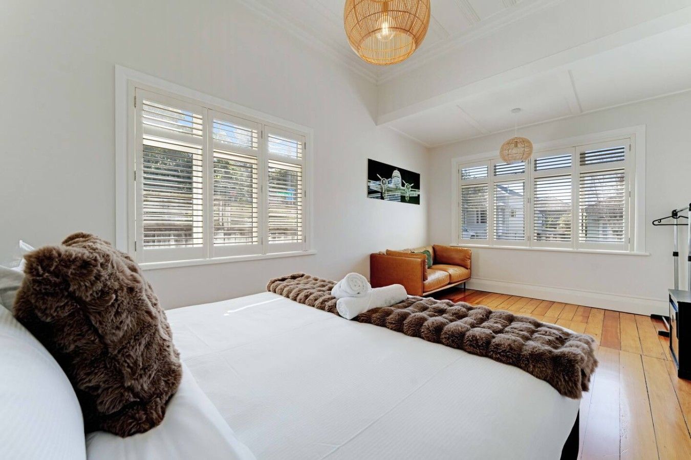 A bedroom with a large bed, two windows with blinds, a brown leather sofa, and a hanging light fixture.