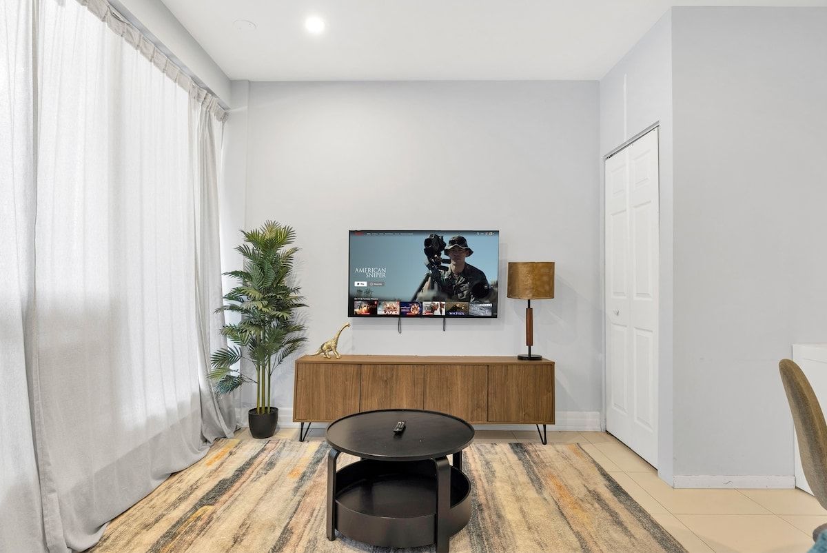A living room with a flat-screen TV mounted on the wall, a round coffee table, a potted plant, and a wooden cabinet.