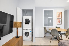 Un espace de vie compact comprenant un téléviseur à écran plat, un lave-linge, un coin repas avec des chaises et une fenêtre agrémentée d’œuvres d’art.