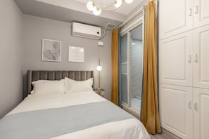 Une chambre d’hôtel comprenant un lit, une table de chevet, un climatiseur fixé au mur, un certificat encadré et une porte-fenêtre coulissante avec des rideaux.