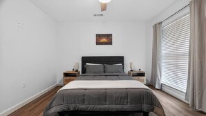 Une chambre à coucher comprenant un lit, des tables de chevet, une commode, un tableau accroché au mur et une fenêtre avec des rideaux.