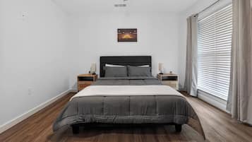 Un dormitorio con cama, mesitas de noche, un cuadro en la pared y una ventana con cortinas.