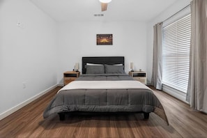 Un dormitorio con cama, mesitas de noche, un cuadro en la pared y una ventana con cortinas.