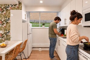 A modern kitchen with white cabinetry, a dining area with a wicker chair, and a view of greenery outside. - Boho 2BR | Monthly Stays | Quiet East Ottawa (Ottawa)