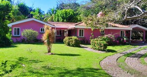 A pink house with a red roof, surrounded by lush greenery and a well-maintained lawn.
