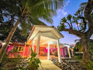 A covered outdoor seating area with a stone pathway leading to a building with a pink facade. - Rancho Colibrí (Acajutla)