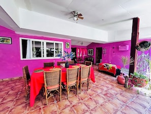 A dining area with a red table and wicker chairs, a ceiling fan, and a pink wall. - Rancho Colibrí (Acajutla)
