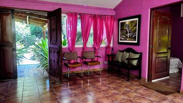 A room with a wooden door, a bench, two chairs, a window with pink curtains, and a framed picture on the wall.