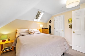 A bedroom with a bed, a nightstand, a lamp, a dresser, a coat hanging, and a door. - Downtown Bozeman Craftsman Near MSU & Main St (Bozeman)