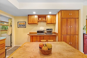 A compact kitchen with wooden cabinets, a microwave, an oven, and a dining table with a fruit bowl. - Downtown Bozeman Craftsman Near MSU & Main St (Bozeman)