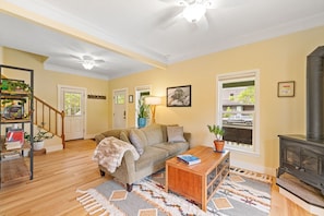 A living room with a sofa, coffee table, and fireplace. There is a staircase with a railing, a door, and a window with a view. - Downtown Bozeman Craftsman Near MSU & Main St (Bozeman)