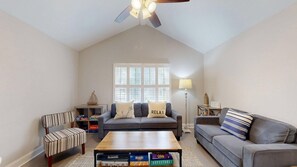 A living room with a high ceiling, a ceiling fan, a window with blinds, a sofa with cushions, a striped armchair, a coffee table with board games, and a lamp. - New to VRBO, 5BR Palmetto Dunes, 7.5 Min Walk To Beach, Private Pool (Hilton Head Island)
