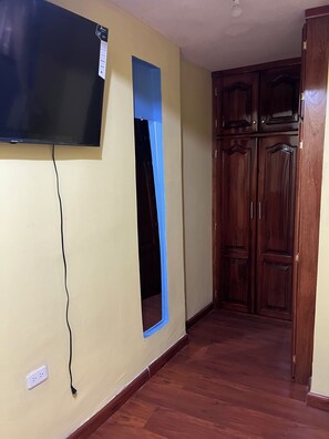 Una habitació amb televisió penjada a la paret, un armari de fusta i una finestra amb marc blau.