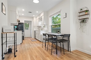 A modern kitchen with white cabinets, a dining area with a table and chairs, and a window with a view of greenery. - F1 Ready - Heart of South Austin - 2B/1.25BA - King Bed - Fast WiFi - Yard (Austin)
