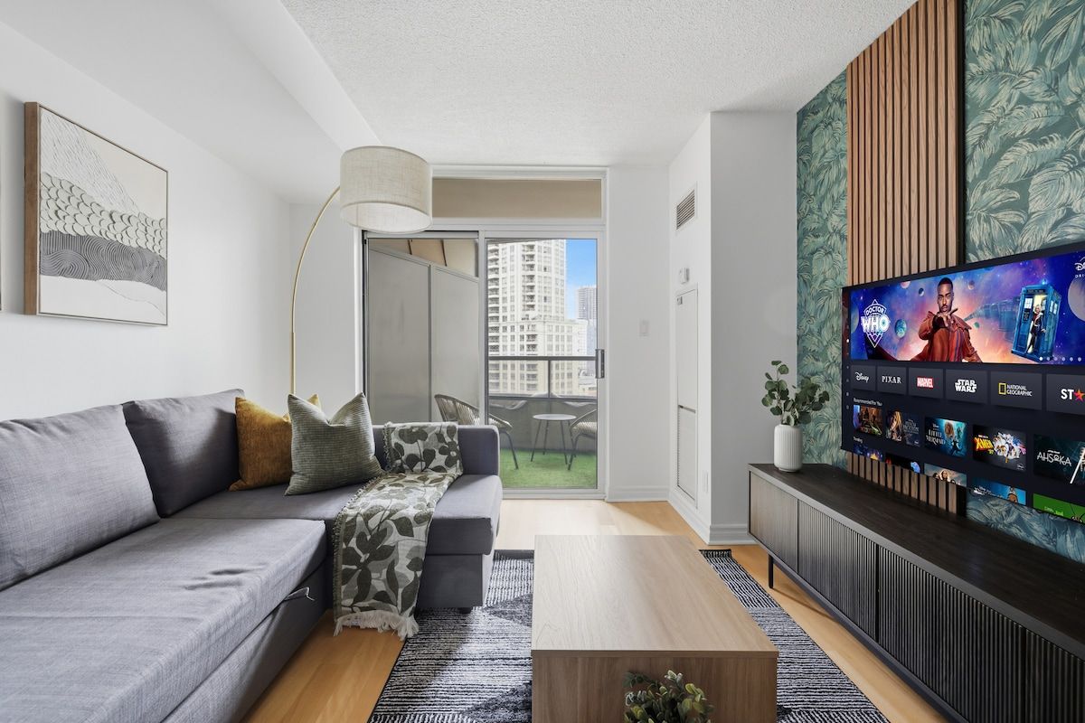 A modern living room with a grey sofa, a coffee table, and a flat-screen TV mounted on the wall displaying a movie selection.