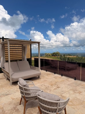 An outdoor seating area with a cushioned sofa and wicker chairs, a pergola with a wooden slatted roof, and a view of the landscape.