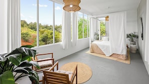 Ein geräumiges Zimmer mit großen Fenstern, einem Bett, einer Sitzecke mit rundem Teppich und einer Pendelleuchte.