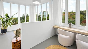 Ein modernes Zimmer mit großen Fenstern, einem Topf mit Pflanze, einem Holznachttisch und einem runden Teppich aus Gewebe.