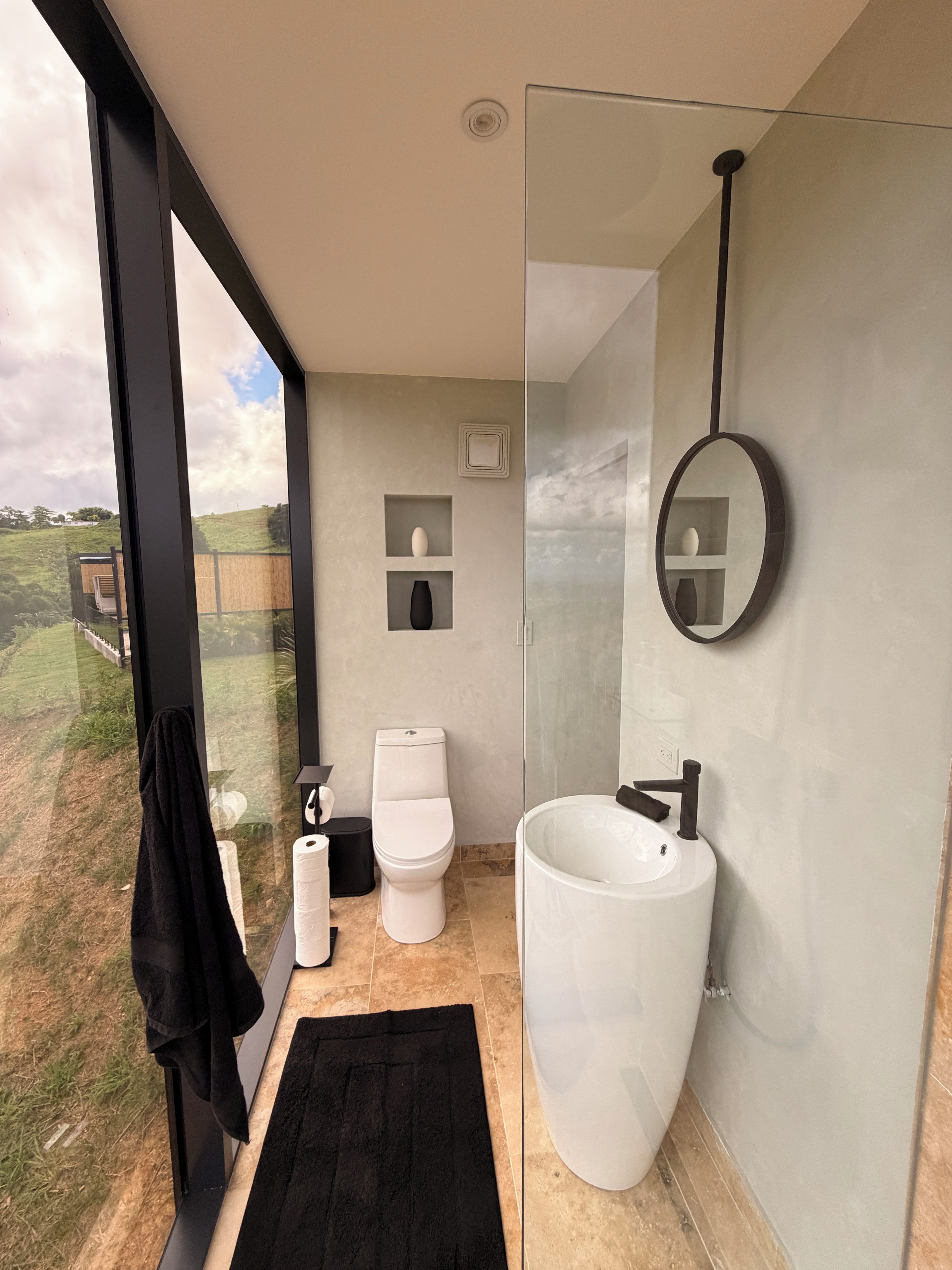 Een moderne badkamer met een groot raam, een wit toilet, een witte wastafel en een zwarte handdoek.