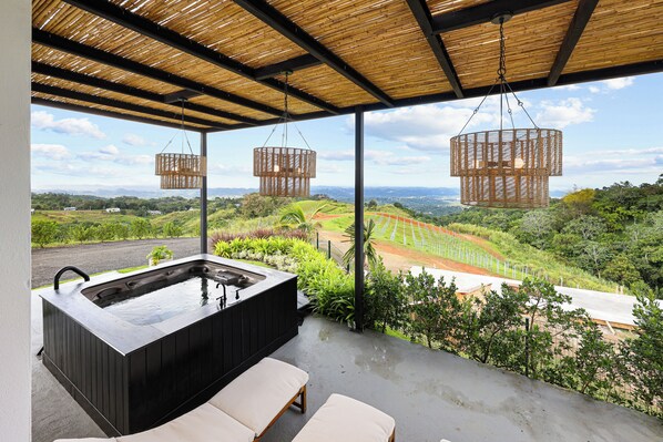 A hot tub with a wooden exterior, two hanging wicker lamps, and a view of a landscaped garden and distant hills.