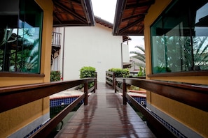 A wooden walkway leading to a pool area with tiled flooring, surrounded by greenery and a building with large windows. - Costa do Sol By MN Hoteis (Búzios)