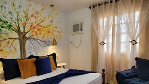 Quarto com cama, cadeira, ar-condicionado na parede e janela com cortinas leves.