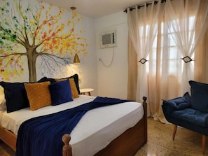 Un dormitorio con cama, una silla, aire acondicionado de pared y una ventana con cortinas translúcidas.
