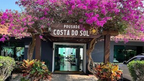 Entrada de hotel com placa indicando 'Pousada Costa do Sol', cercada por plantas em vasos e um carro estacionado nas proximidades.