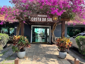 Entrada de hotel con un letrero que dice 'Pousada Costa do Sol', rodeada de plantas en macetas y un coche estacionado cerca.