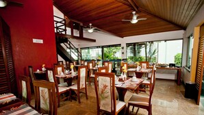 Um restaurante com várias mesas e cadeiras, uma escadaria e grandes janelas.