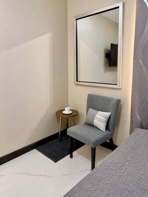 Una habitación de hotel con un sillón gris, una mesita redonda con una taza blanca, un espejo grande y una colcha estampada.