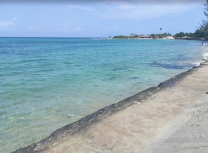 A beach with clear turquoise water, a sandy shore, and a distant view of a residential area with buildings and vegetation. - Escape to Paradise!| Your Home Away From Home in Jamaica (Runaway bay)