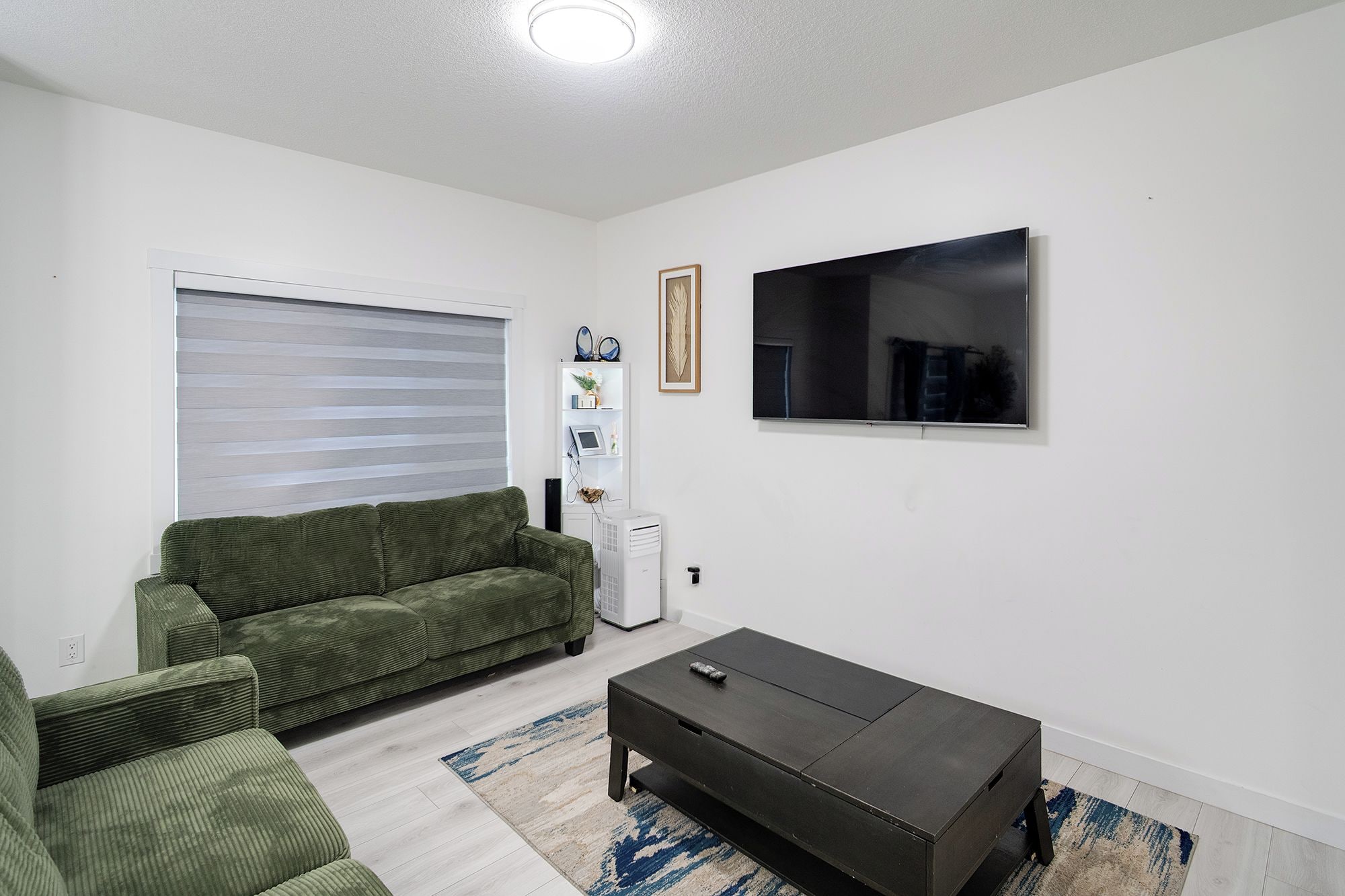 A living room with a green sofa, a black coffee table, a flat-screen TV, and a window with blinds.