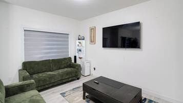 A living room with a green sofa, a black coffee table, a flat-screen TV, and a window with blinds.