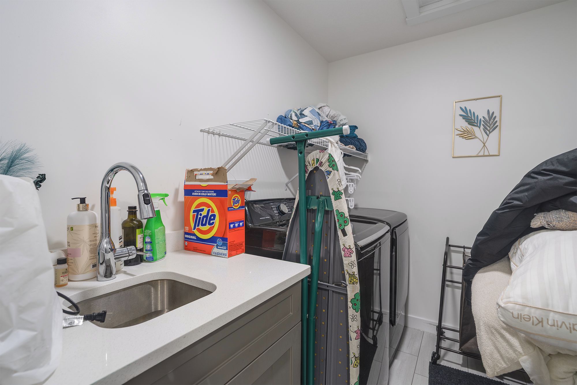 A compact kitchen with a sink, a drying rack, and a box of Tide detergent.