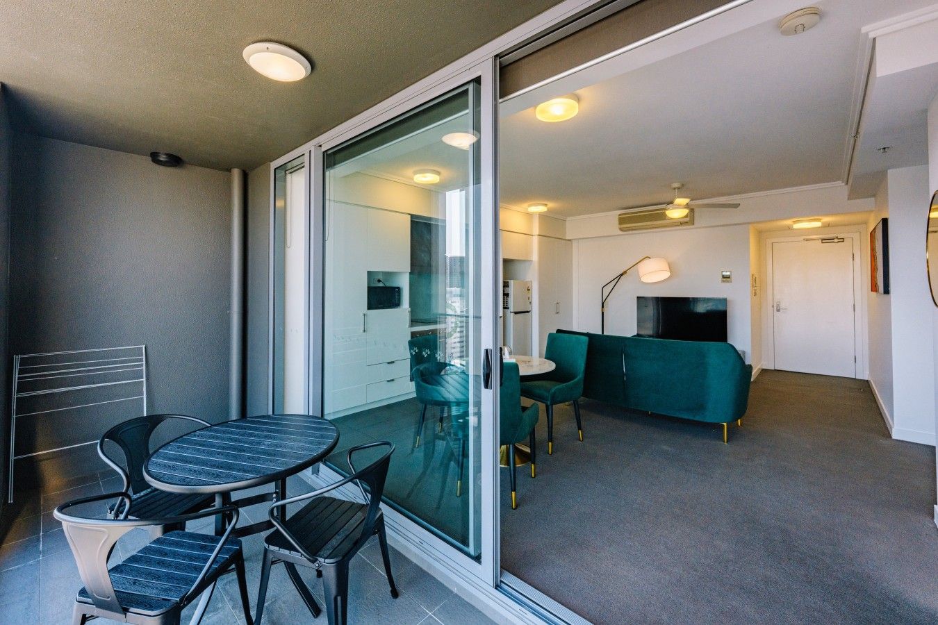 A modern living room with a glass door leading to a balcony, a green sofa, a TV, and a dining area with a table and chairs.