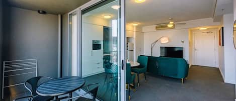 A modern living room with a glass door leading to a balcony, a green sofa, a TV, and a dining area with a table and chairs.