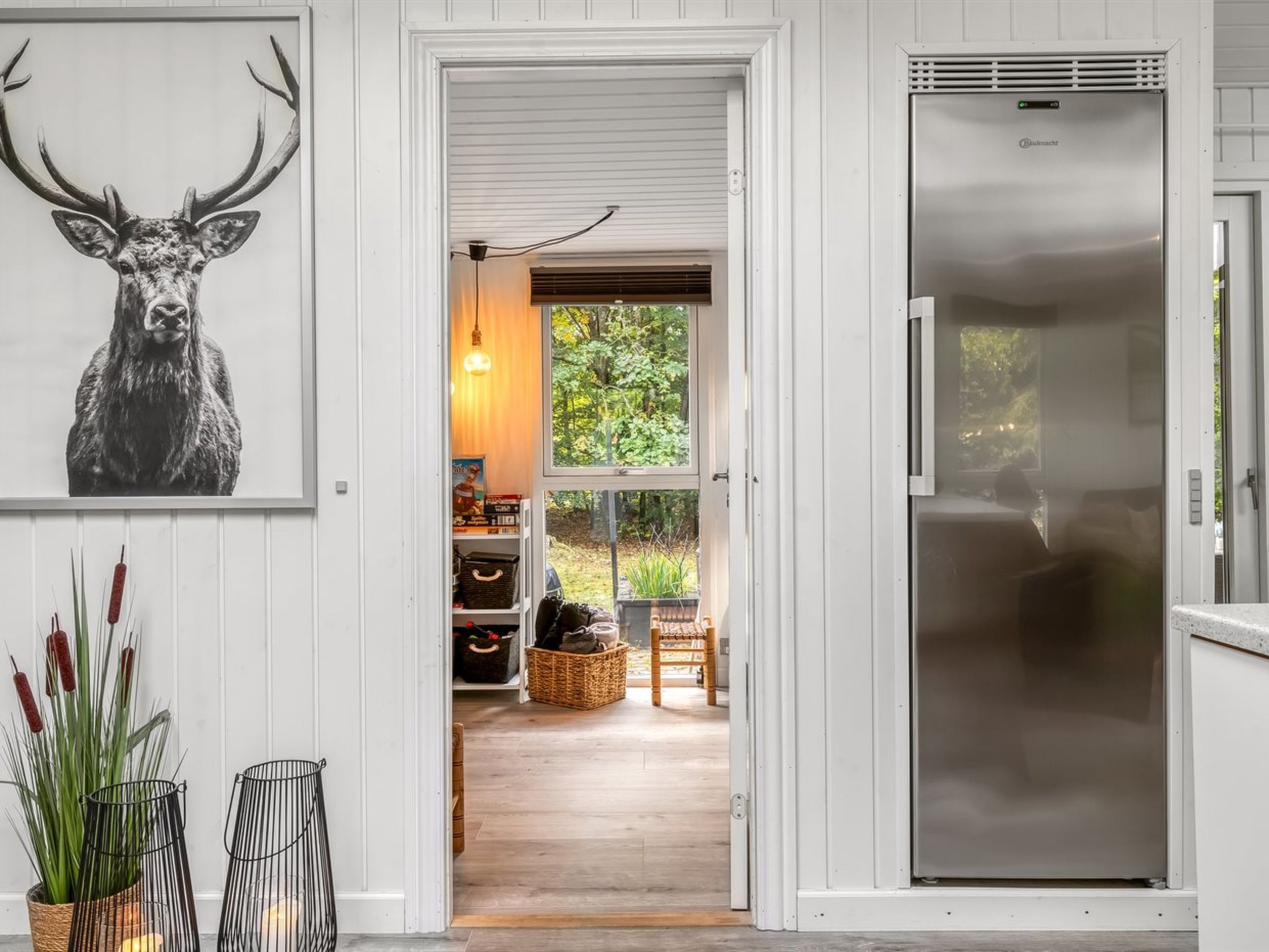 A modern kitchen with a large stainless steel refrigerator, a wooden floor, and a view through an open door to a dining area with a table and chairs.