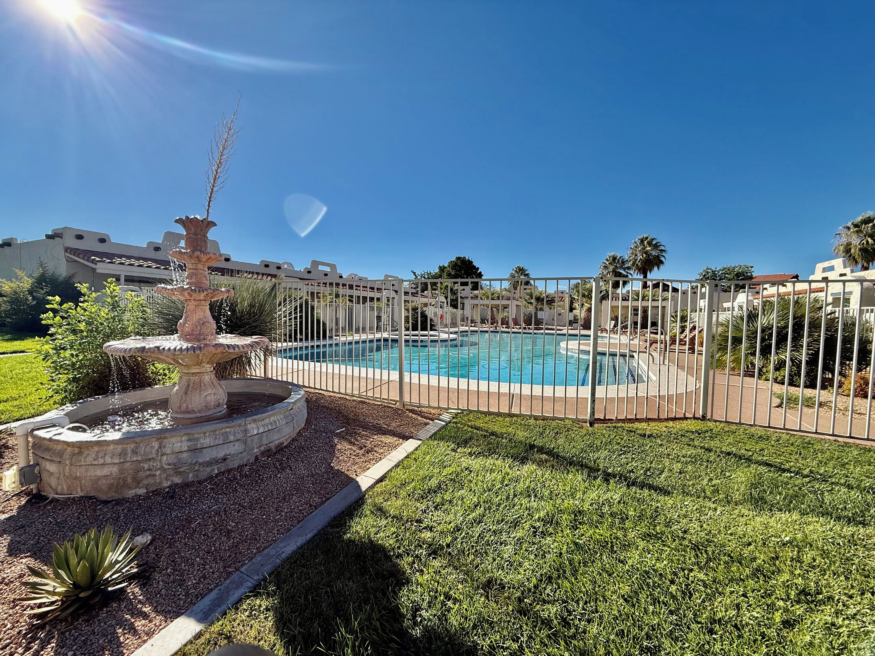A swimming pool with a fountain, surrounded by a fence, and a grassy area with a small garden.