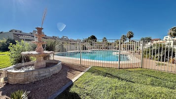 A swimming pool with a fountain, surrounded by a fence, and a grassy area with a small garden.