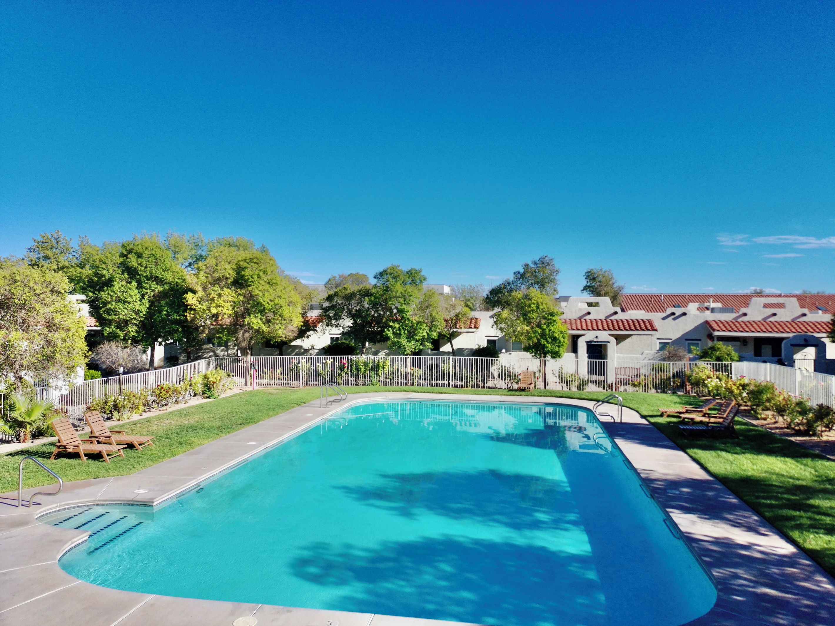 Une piscine rectangulaire à l’eau d’un bleu limpide, entourée d’une clôture blanche, et une zone herbeuse agrémentée d’arbres.