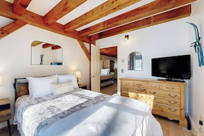 A bedroom with a large bed, wooden ceiling beams, a dresser, a television, and a mirror. - Riverbend Ski Retreat - Modern Condo minutes to Sunday River, Breweries & Bethel (Bethel)