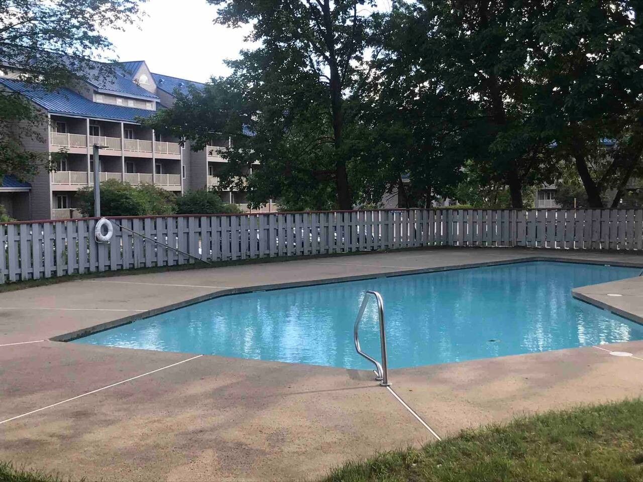 A swimming pool with a metal ladder, surrounded by a wooden fence and a building with balconies in the background.