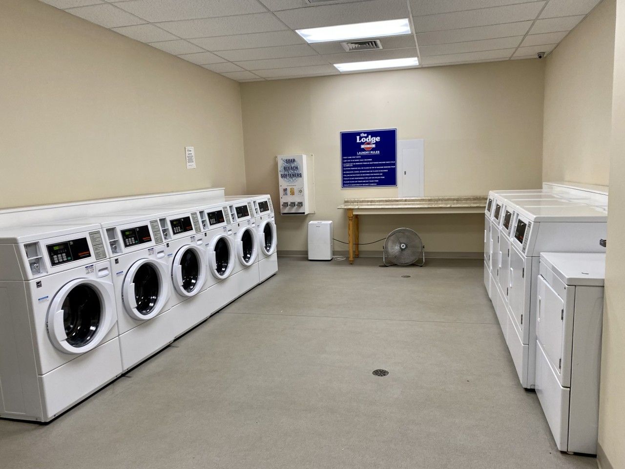 A laundry facility with multiple washing machines and dryers, a fan, and a sign with laundry rules.