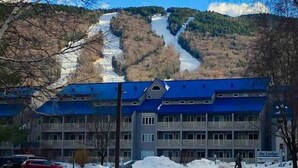 Um edifício com telhado azul, um estacionamento com neve e uma montanha ao fundo.