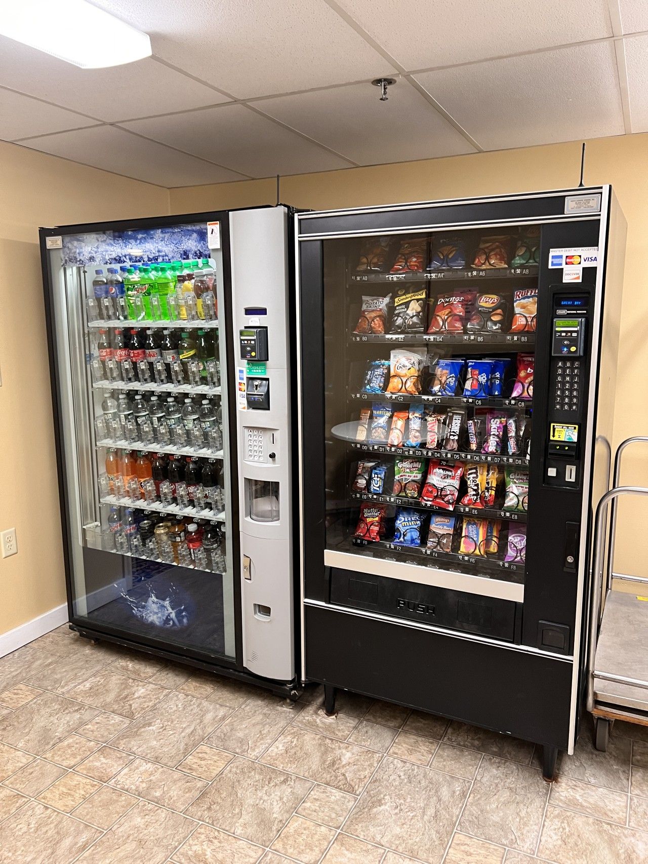 Two vending machines in a room with beige walls and tiled flooring.