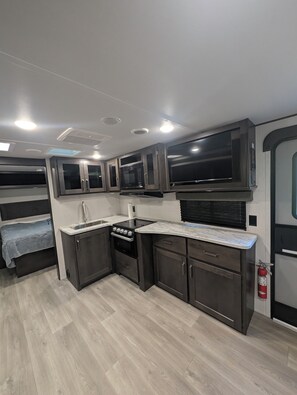 Een compacte keuken met een fornuis, magnetron en kasten, grenzend aan een slaapgedeelte met een bed.