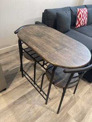 Mesa de jantar com tampo de madeira e estrutura metálica, em um quarto com sofá cinza e almofada estampada.